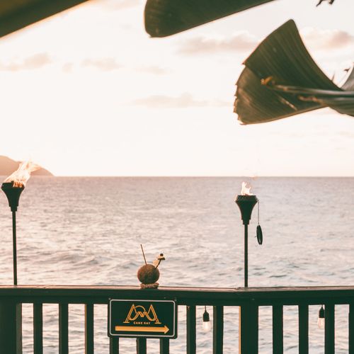 A tropical seascape with tiki torches on a wooden railing, overlooking the ocean at sunset. A drink is placed on the railing.