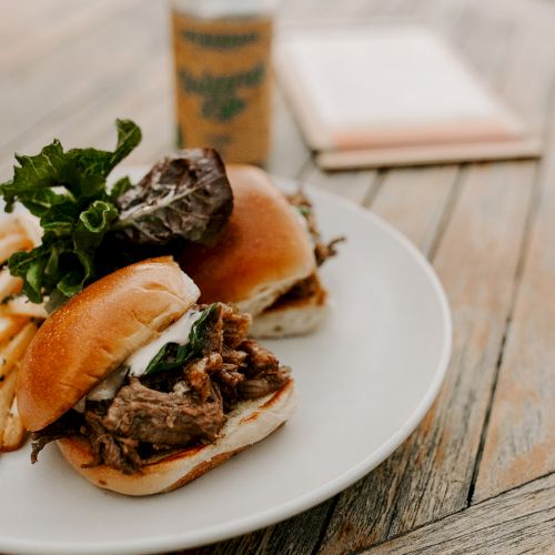 A plate with two sliders filled with pulled meat and greens, served with a side of fries on a wooden table.