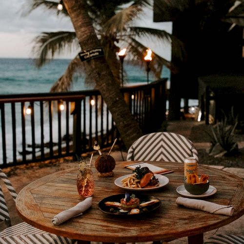 A seaside table with drinks and food, surrounded by chairs, set against an ocean backdrop with palm trees and warm lighting.
