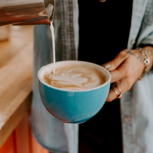 A person is pouring milk into a latte in a blue cup, creating latte art. They are wearing a denim shirt and holding the cup carefully.