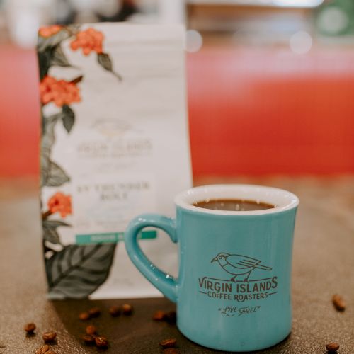 A blue mug filled with coffee is placed in front of a coffee bag; coffee beans are scattered around the table.