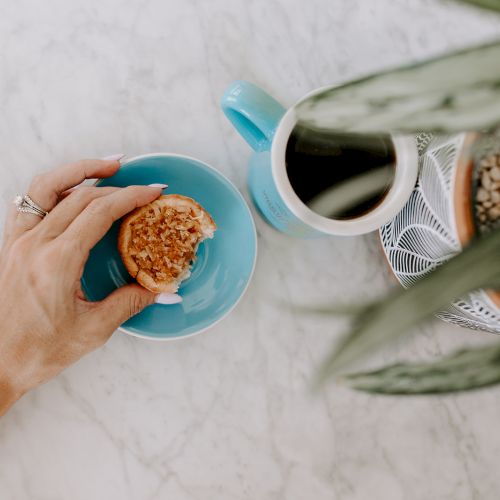 A hand holding a cookie on a blue plate near a mug of coffee, with a potted plant on a marble surface.