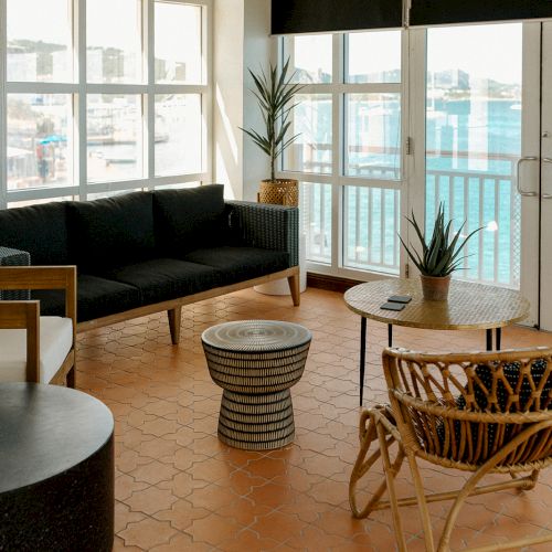 A cozy room with seating, wooden furniture, and a view of the sea through large windows. The floor is tiled in a warm color.