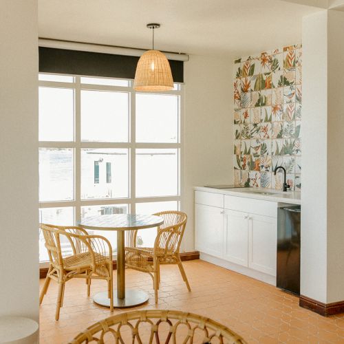The image shows a bright room with a glass table, wicker chairs, large windows, and a small kitchenette with a colorful tile backsplash.