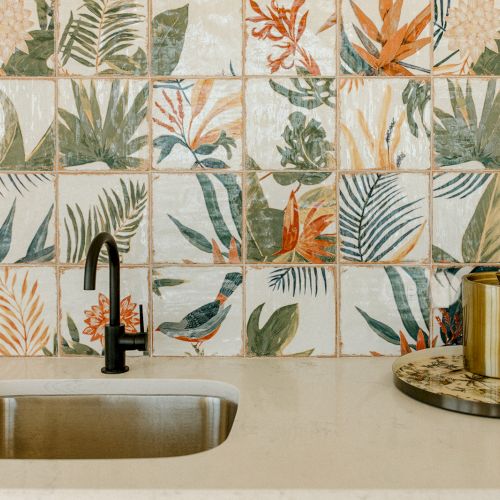 A kitchen sink with a black faucet is set against floral-patterned tiles, with a decorative tray and candle on the countertop.