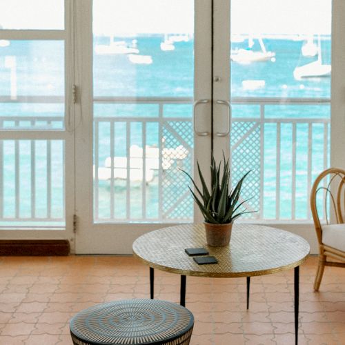A cozy room with wicker furniture, a table with a plant and a phone, facing large windows showing a view of the sea and boats outside.