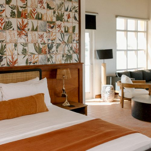 A stylish room with a large bed, tropical-themed wall art, sofa, coffee table, and large windows letting in natural light.