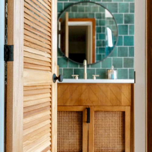 A partially open wooden door reveals a bathroom with a circular mirror, green tiles, and a wooden vanity with woven texture cabinetry.