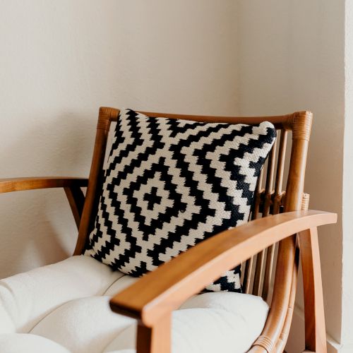 A wooden chair with a white cushion and a black-and-white geometric patterned pillow in a corner space.