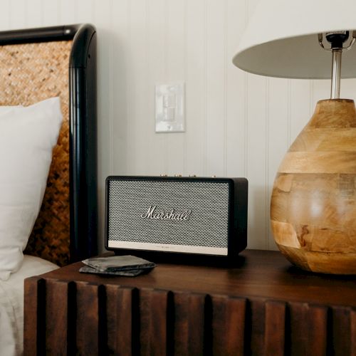 A bedside setup with a pillow, wooden lamp, speaker, and phone on a rustic nightstand.