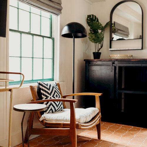 A cozy room with a wooden chair, side table, standing lamp, and large mirror, featuring a potted plant and soft lighting.