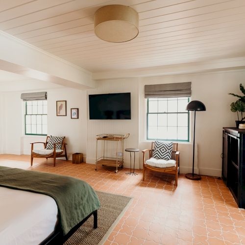 A cozy bedroom with a bed, armchairs, a TV, and décor. The room features a green accent door and earthy tones, with natural light streaming in.