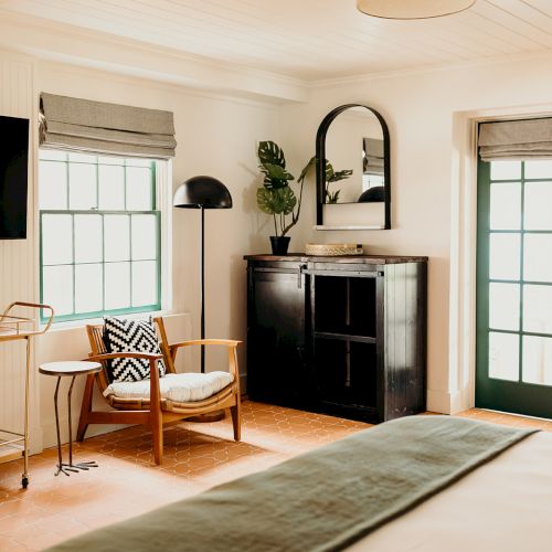 The image shows a cozy room with a bed, armchair, dresser, TV, potted plant, and a trolley. Sunlight filters through the windows.