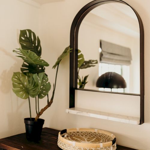 A room with a dark wood table, black potted plant, woven basket, and arched mirror reflecting a window and lamp.