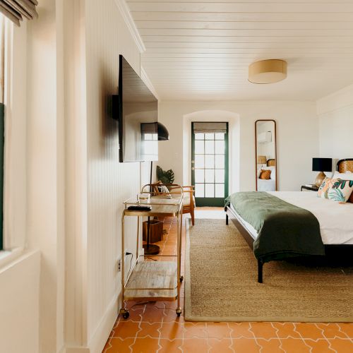 A cozy bedroom with a bed, TV, mirror, and a green blanket. There's a window to the left, a wooden cart, and carpet flooring.