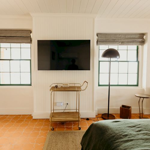 The image shows a cozy room with a TV, two windows, an armchair, a floor lamp, a small table, and a cart on a tiled floor.