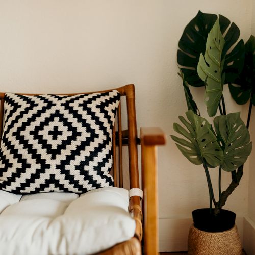A wooden chair with a black-and-white patterned pillow sits next to a potted plant with large green leaves in a cozy corner.