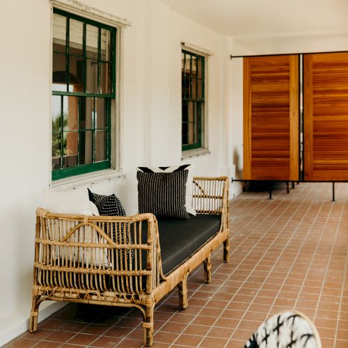 A narrow corridor with a wicker couch, black cushions, tiled floor, green-trimmed windows, and wooden sliding doors at the end.