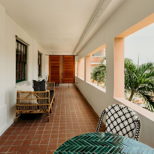 A sunlit balcony with wicker seating, patterned chairs, and a green table overlooks a tropical landscape and a body of water.