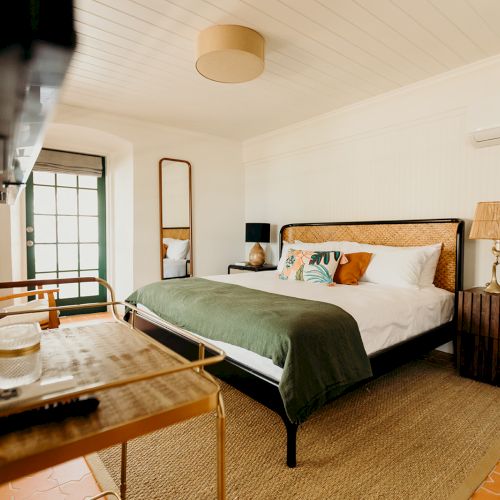 A cozy bedroom with a large bed, green throw, side table, decor items, and a TV on the left wall, exuding a warm and inviting vibe.