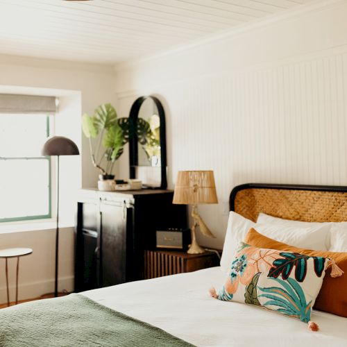 A cozy bedroom features a large bed with decorative pillows, a side table, a lamp, and a chair by the window with plants placed nearby.