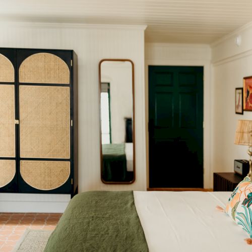 A stylish bedroom featuring a rattan wardrobe, a large mirror, and a neatly made bed with colorful pillows and a green blanket.