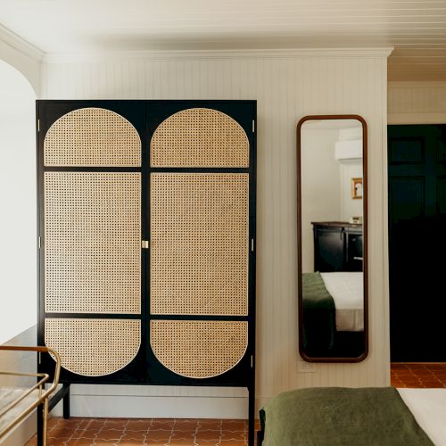 The image shows a room with a wardrobe, a floor mirror, a bed with a green blanket, and a rug on terracotta tiles.