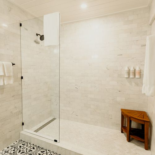 A clean, modern shower with marble walls, a glass door, black fixtures, wooden stool, and neatly hung towels.