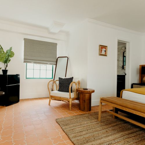 A cozy bedroom with a bed, woven chair, plants, wall art, and a mirror on terracotta-tiled flooring.