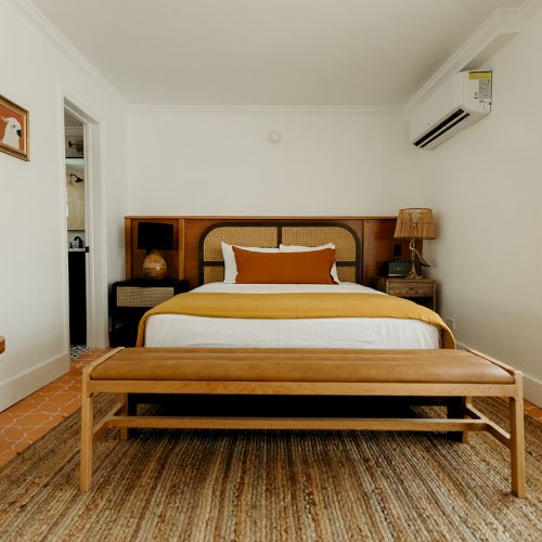 A cozy bedroom with a wooden bed, bench, side tables, and a window. The room has neutral colors and a warm, inviting atmosphere.