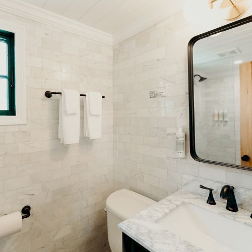 A modern bathroom with a white tiled wall, a large mirror, a sink with black fixtures, and a toilet.