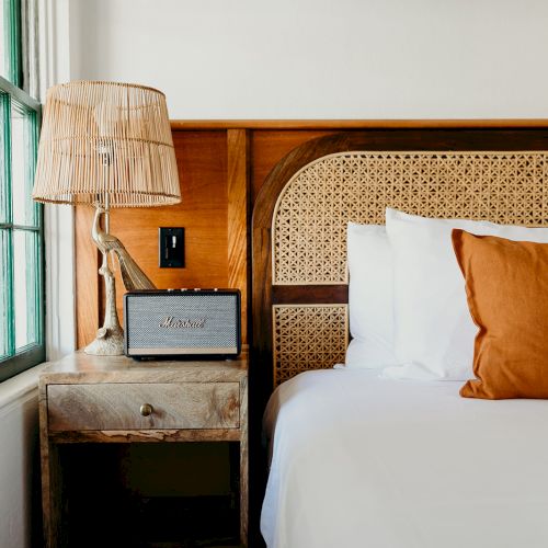 The image shows a cozy bedroom corner with a bed, orange pillow, rustic nightstand, lamp, and small radio by a window.