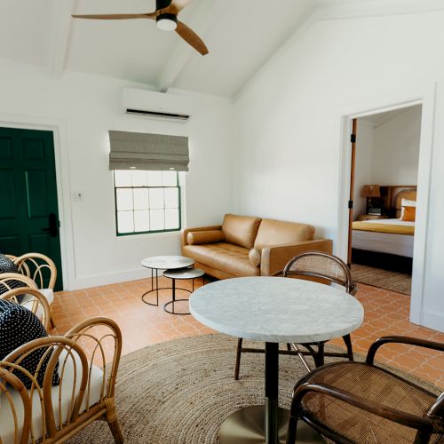 A cozy living room with rattan chairs, a sofa, tables, a rug, and a plant; a bedroom is visible through the open doorway.