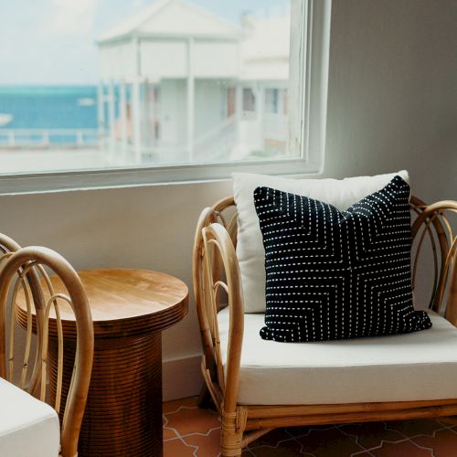 A cozy seating area with wicker chairs, a side table, and a view of the ocean through a window. A black patterned pillow adds contrast.
