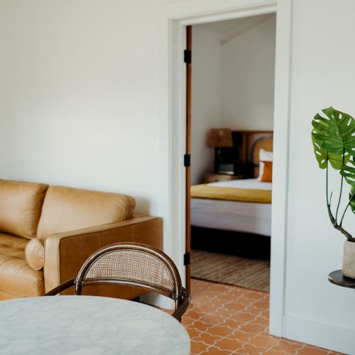 The image shows a cozy living room with a brown sofa, a table, a chair, and a potted plant, leading into a bedroom with a bed.