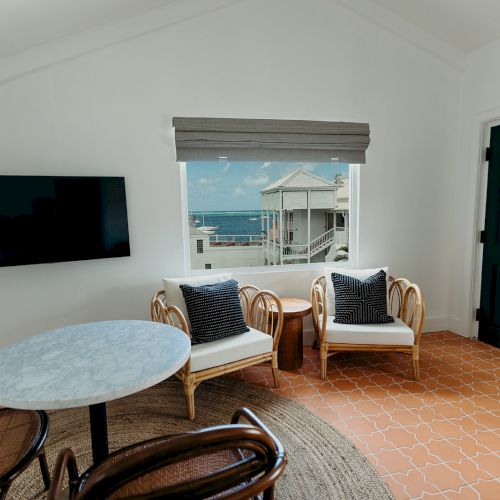 A cozy room with chairs, a round table, a wall-mounted TV, and a window view of buildings and the ocean.