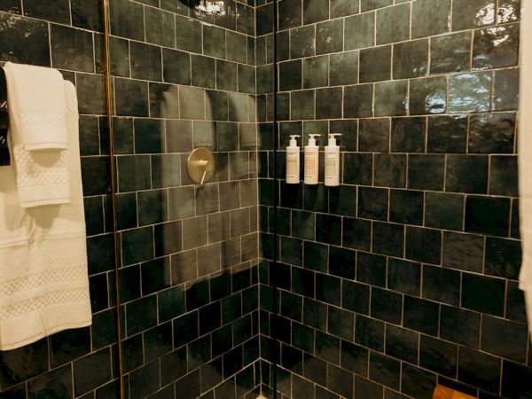 A modern shower with dark tiles, glass door, wooden stool, and bottles on the wall. Towels are hung nearby.