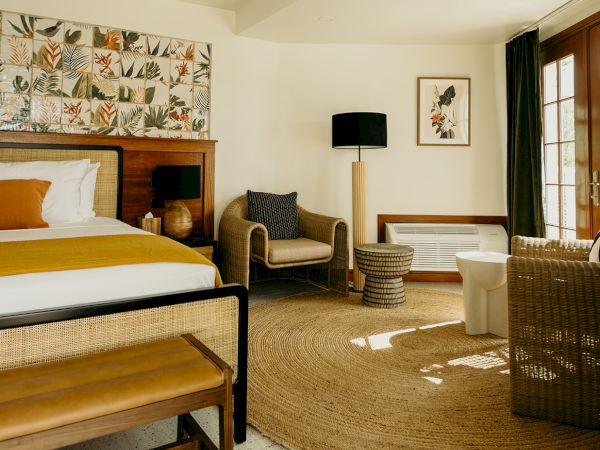 A stylish bedroom features a bed, armchair, lamp, artwork, and a round rug. Sunlight streams through a door beside a small seating area.