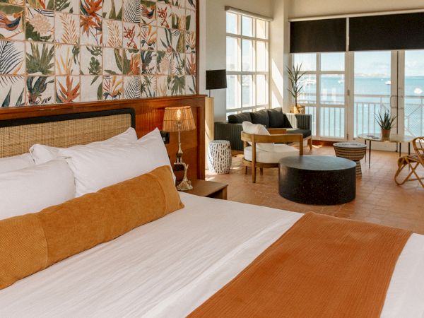 A cozy hotel room with a large bed, tropical decor, and a sitting area overlooks a balcony with a view of the sea.