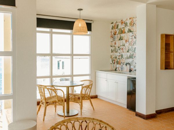 A bright room features a table with four wicker chairs, large windows, a small counter with tiles, and a hanging light fixture.