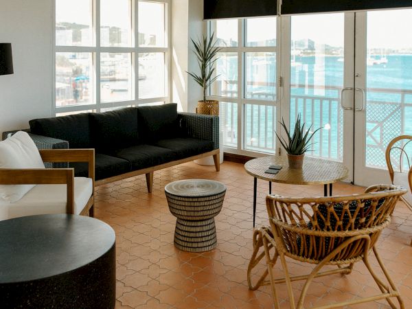 A stylish living room with wicker chairs, a black sofa, and ocean view windows. Coastal decor and a terracotta tile floor complete the scene.