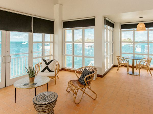 A sunlit room with wicker furniture, large windows, and ocean views. There's an inviting seating area and a small table by the window.