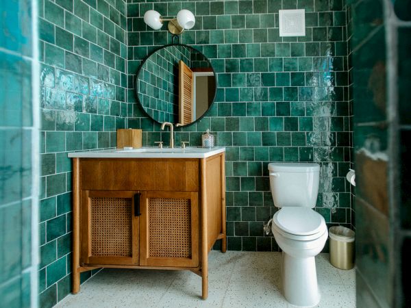 The image shows a bathroom with green tiled walls, a wooden vanity, a round mirror, a toilet, and a dual-light fixture above the mirror.