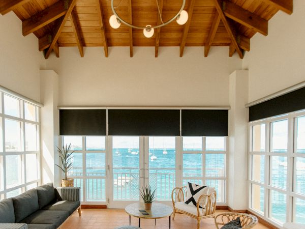 A coastal living room with large windows, a couch, chairs, and a view of the sea. The ceiling is wooden with a modern chandelier.