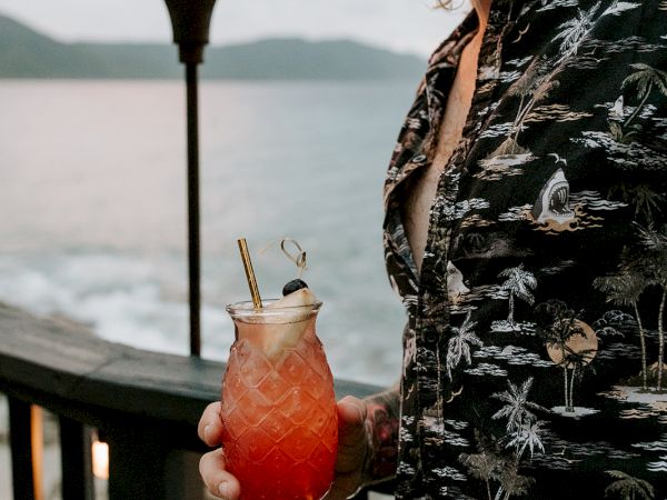A person in a patterned shirt holds a tropical drink by the ocean, with a tiki torch lit in the background during sunset.