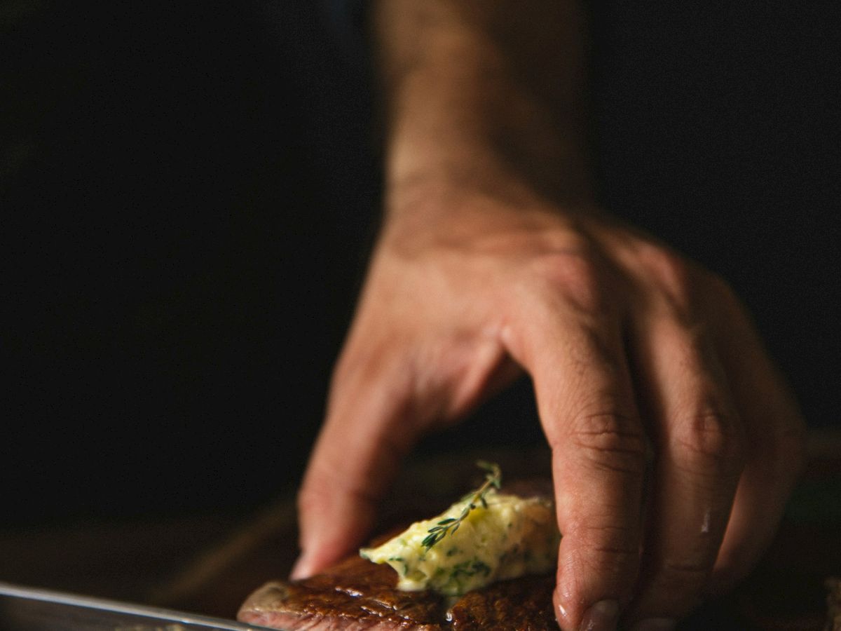 A hand cutting a piece of steak with herb butter on top on a wooden board.