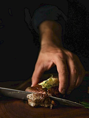 A person slices a steak topped with seasoned butter. The scene is dimly lit, emphasizing the hand and the dish.