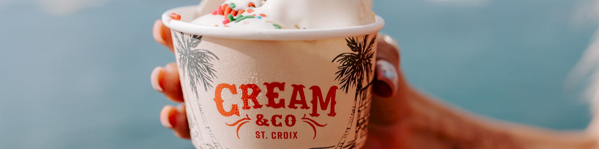 A hand holds a cup of ice cream with sprinkles, labeled "Cream & Co St. Croix," against a scenic waterfront backdrop.