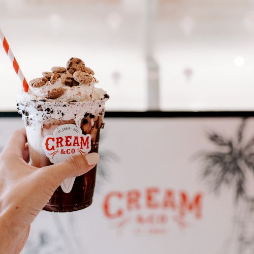 A hand holding a creamy milkshake topped with cookies, whipped cream, and a striped straw, branded with "CREAM & CO".