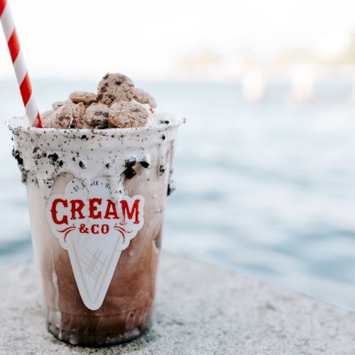 A creamy dessert topped with cookies, in a clear cup labeled "Cream & Co," and a red-striped straw near a waterfront.